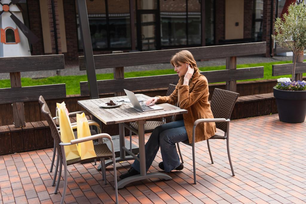 Oldalnézet elegáns nő fülhallgató segítségével laptop közelében kávé és bevásárló táskák teraszon kávézó  - Fotó, kép