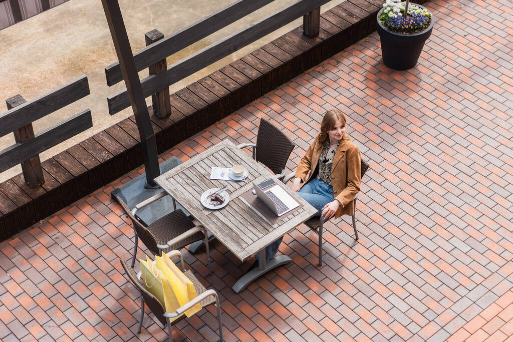 Vista aérea de la mujer con estilo sentado cerca de gadgets, postre y bolsas de compras en la terraza  - Foto, imagen