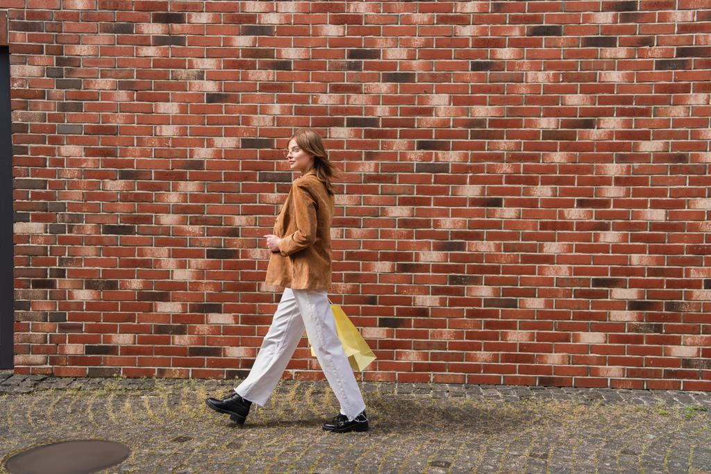 full length of young stylish woman in jacket walking with shopping bags near brick wall  - Photo, Image