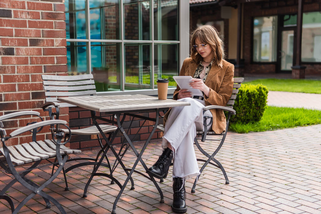 comprimento total da mulher elegante em óculos de sol e jaqueta segurando tablet digital perto de copo de papel na mesa  - Foto, Imagem