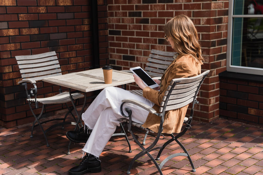 полная длина модной женщины в солнцезащитных очках и пиджаке, держащей цифровой планшет с чистым экраном рядом с бумажной чашкой на столе  - Фото, изображение