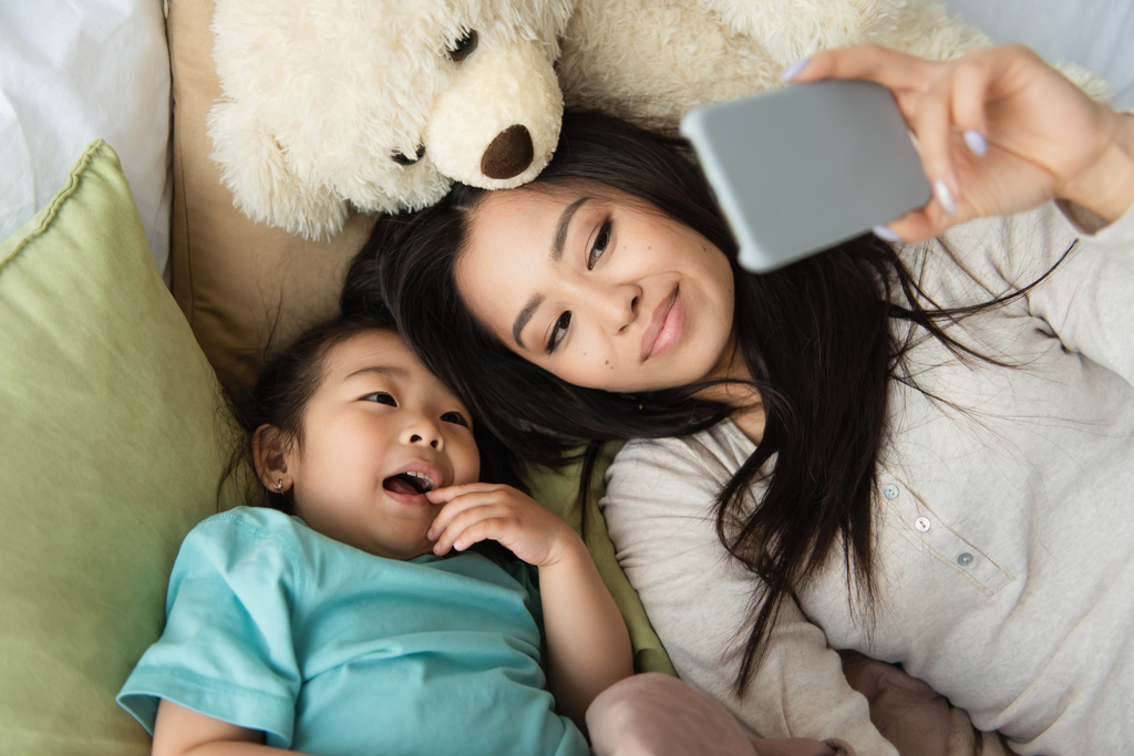 Hoge hoek uitzicht van moeder en aziatische kind het nemen van selfie op smartphone in de buurt teddy beer op bed  - Foto, afbeelding