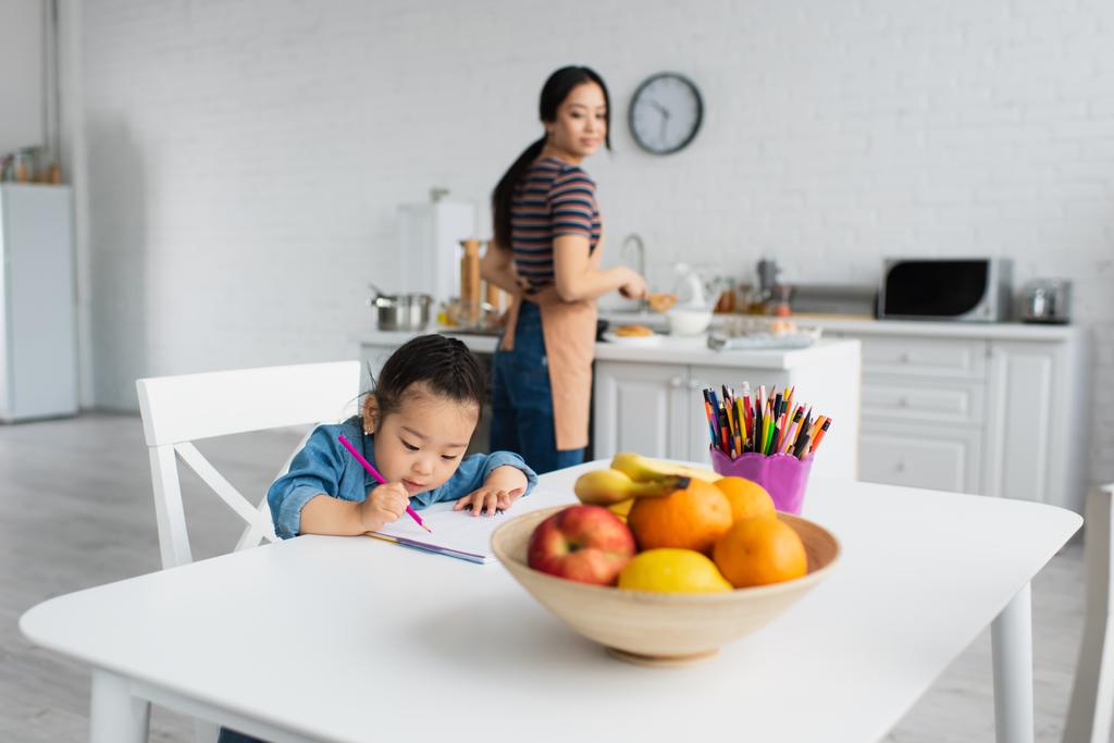 Asiatico bambino disegno vicino frutta e offuscata madre a casa  - Foto, immagini