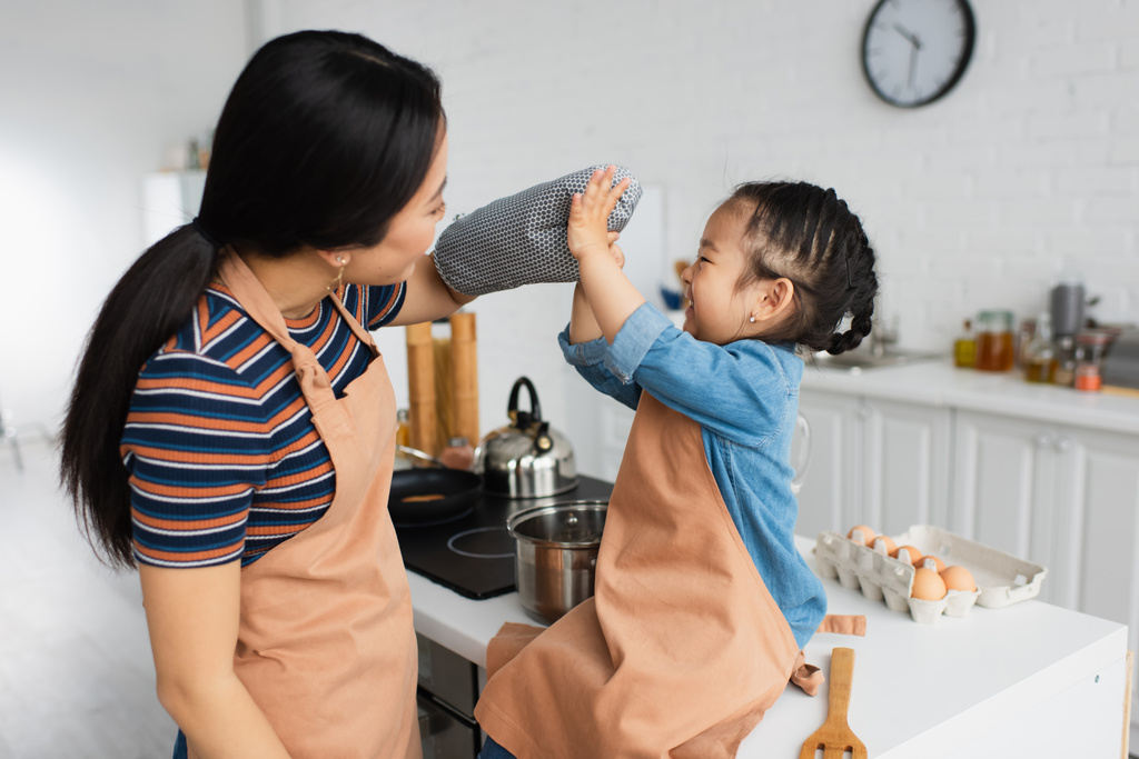 Vista lateral de alegre chica asiática jugando con la madre en el guante del horno en la cocina  - Foto, imagen