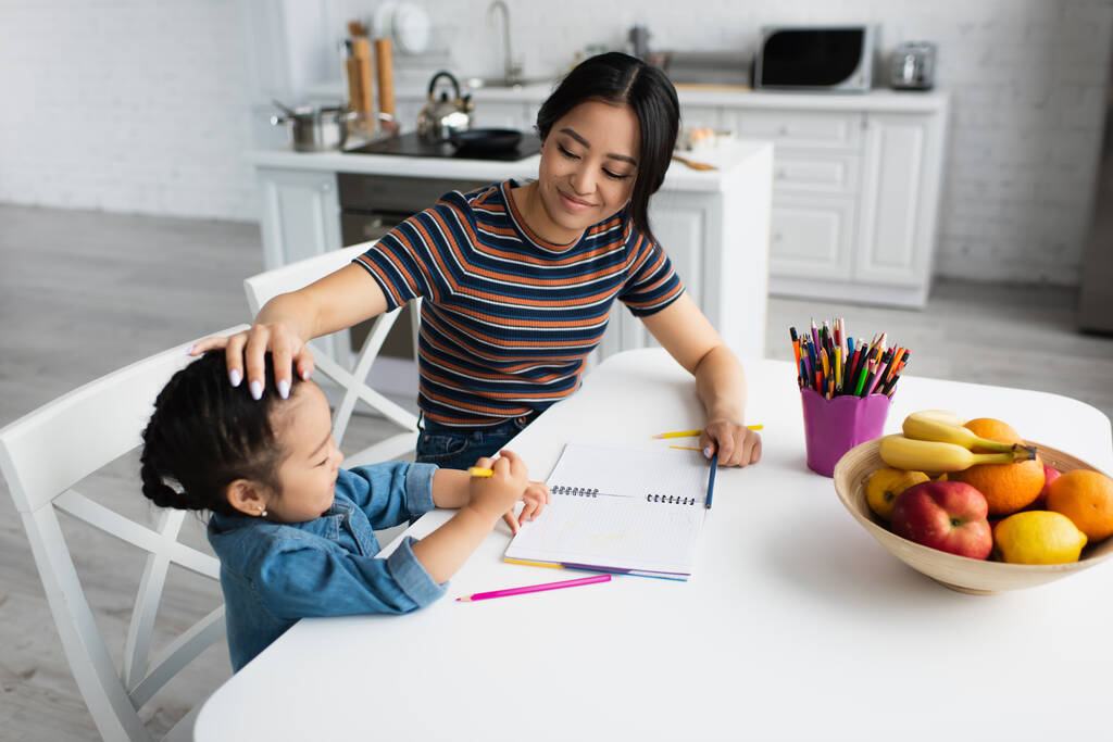 Улыбающаяся мать трогает азиатскую дочь возле цветных карандашей и фруктов на кухне  - Фото, изображение