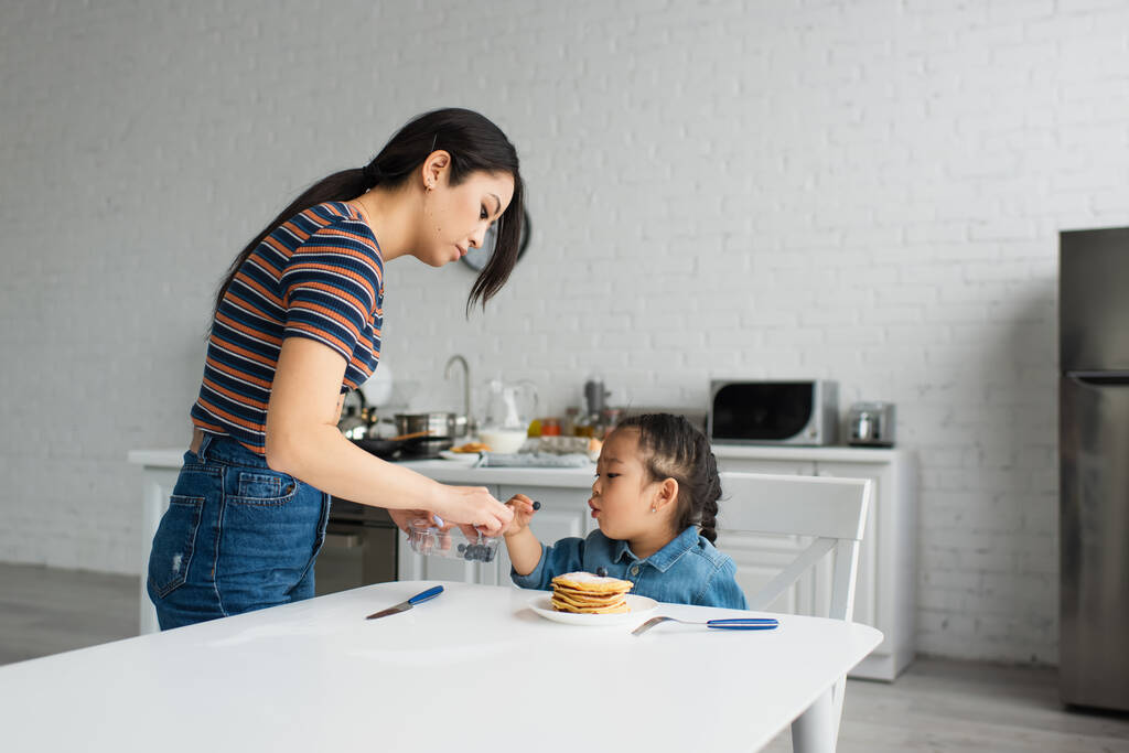 Mère asiatique tenant des bleuets près de l'enfant et des crêpes dans la cuisine  - Photo, image