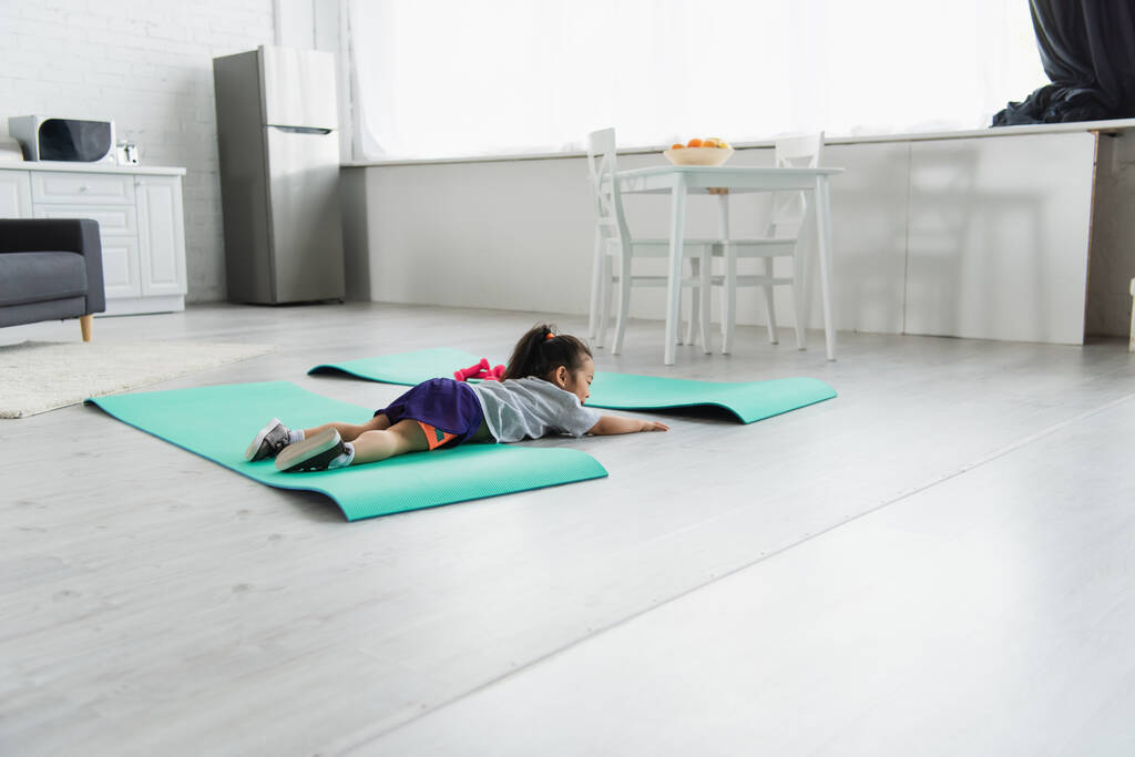 Asiático criança deitado no tapete de fitness perto halteres em casa  - Foto, Imagem