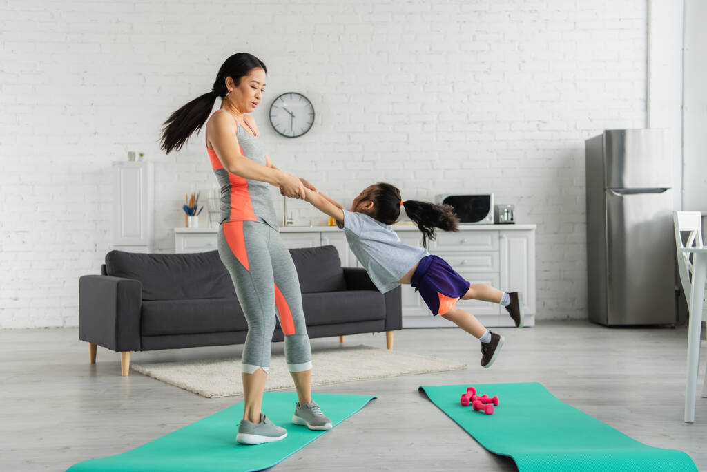 Asian woman spinning daughter near fitness mats at home  - Photo, Image