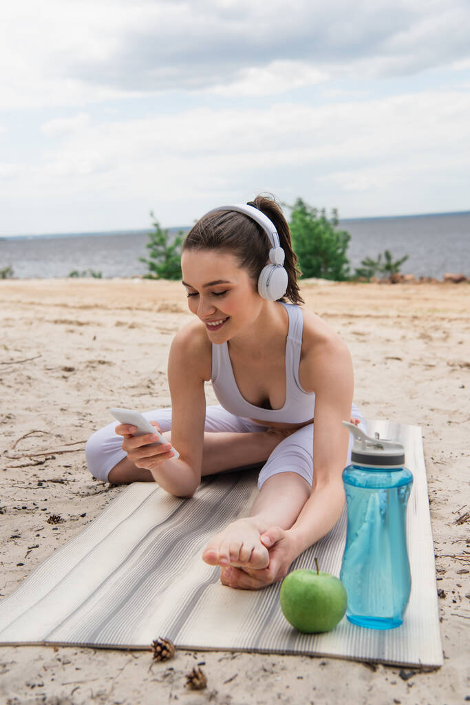 donna sorridente in cuffie wireless ascoltare musica mentre si estende su tappetino yoga e utilizzando smartphone  - Foto, immagini