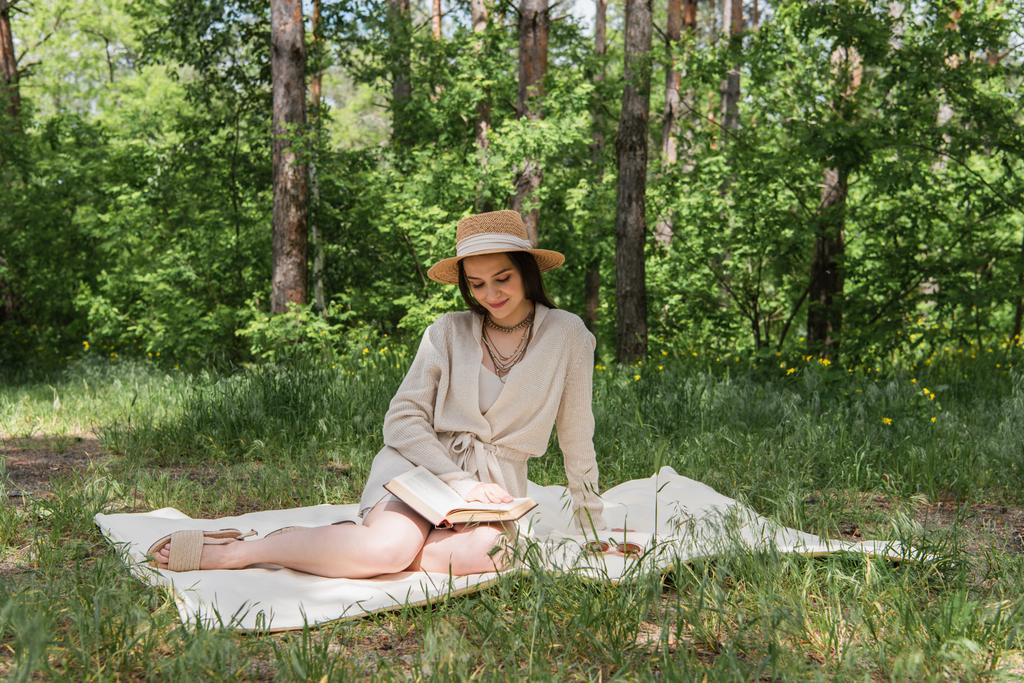 счастливая молодая женщина в соломенной шляпе читает книгу в лесу  - Фото, изображение