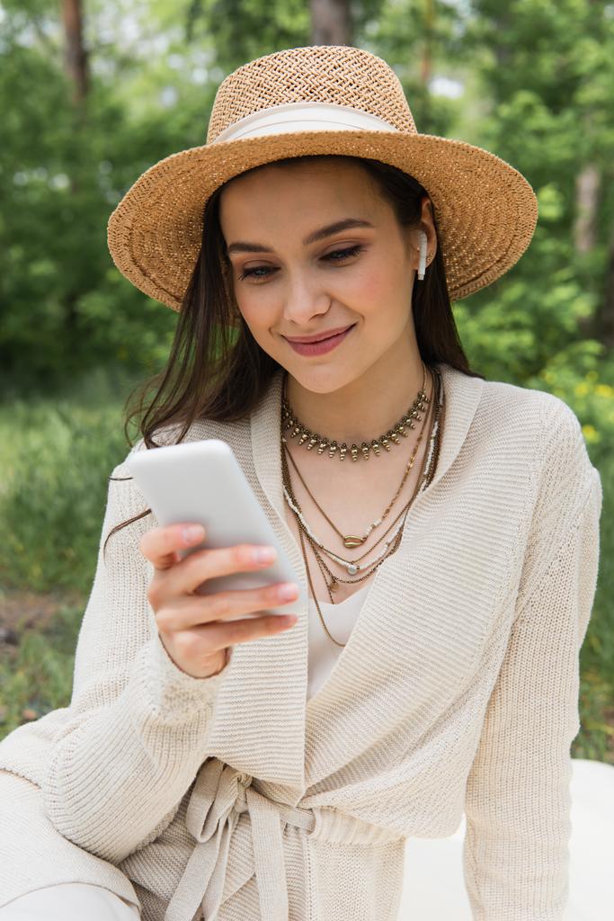 happy young woman in straw hat and wireless earphones using blurred smartphone in forest  - Photo, Image