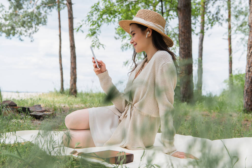 Lächelnde Frau mit Strohhut und drahtlosen Kopfhörern per Smartphone auf Picknickdecke  - Foto, Bild