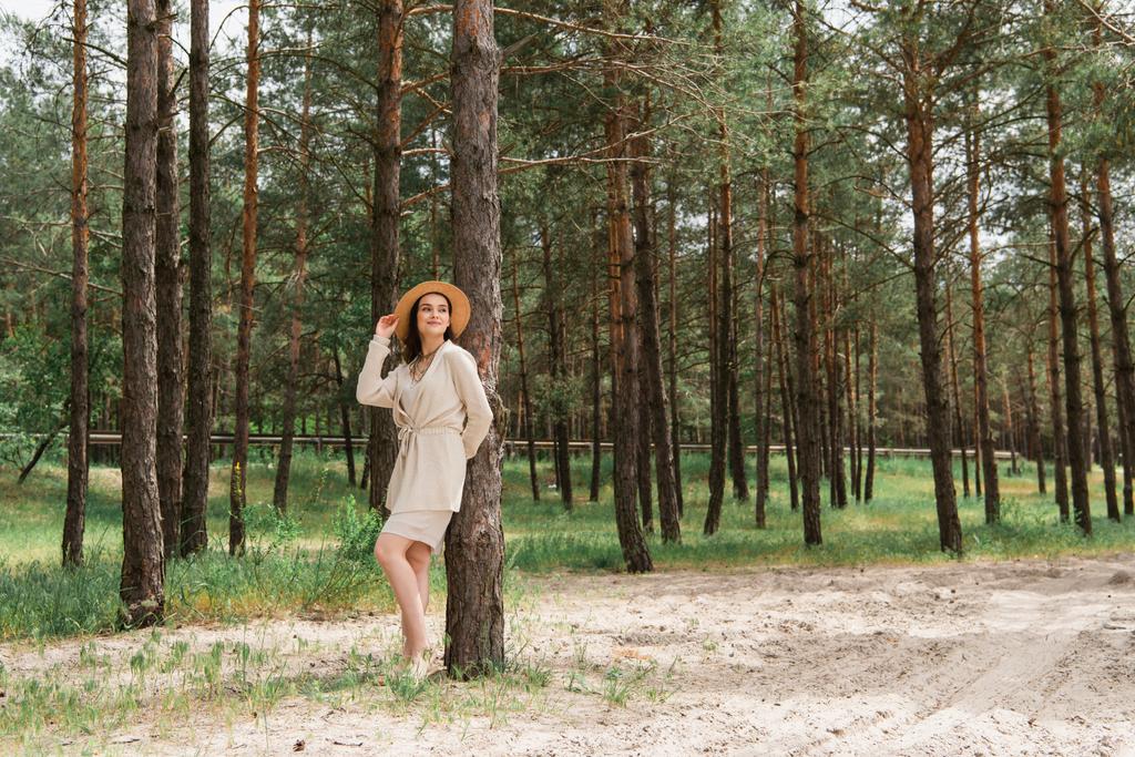 longitud completa de alegre joven mujer ajustando sombrero de paja y de pie en el bosque - Foto, imagen