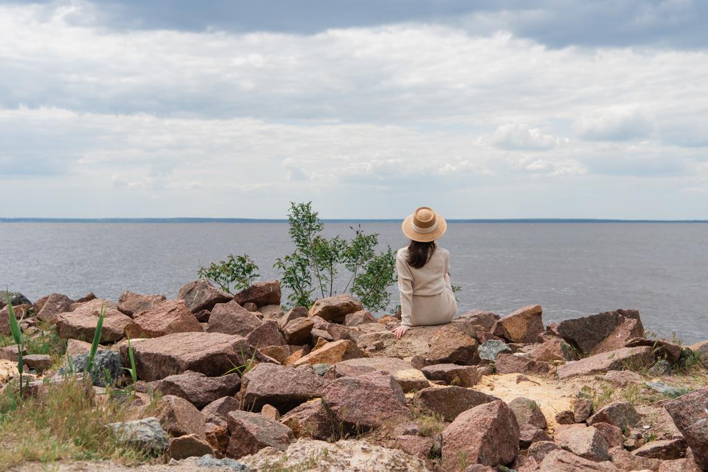 πίσω όψη της νεαρής γυναίκας σε ψάθινο καπέλο κάθεται κοντά στη θάλασσα - Φωτογραφία, εικόνα