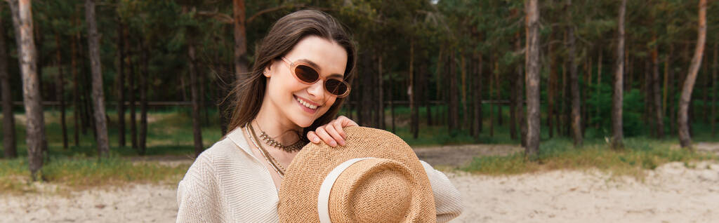 joyeux jeune femme dans les lunettes de soleil tenant chapeau de paille près de la forêt, bannière - Photo, image