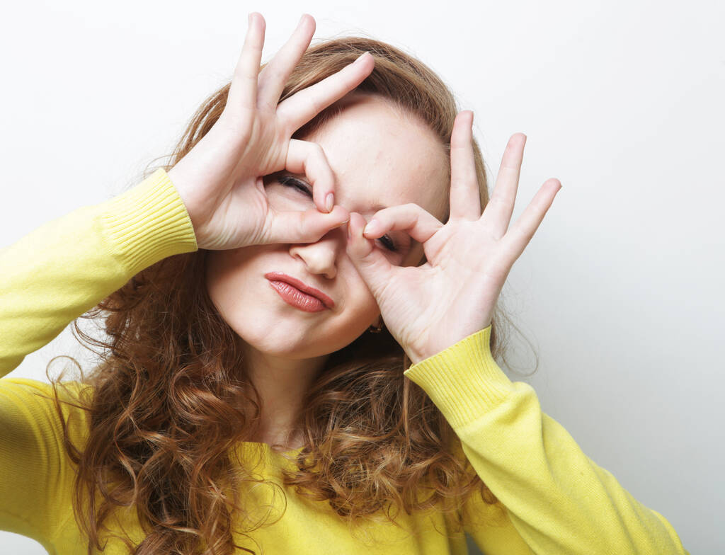 Wesoły kręcone kobieta patrzy przez zero lub ok gest, trzyma zaokrąglone palce w pobliżu oka - Zdjęcie, obraz