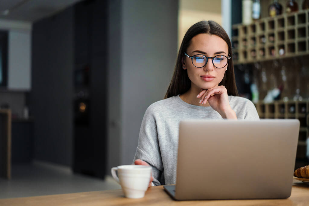 Сосредоточенная молодая женщина в очках пьет кофе во время работы с ноутбуком дома - Фото, изображение