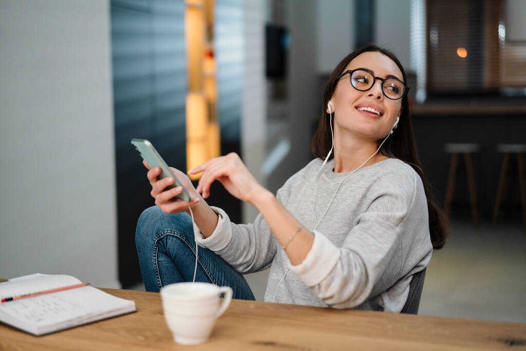 Mujer joven y feliz escuchando música con auriculares y teléfono móvil mientras está sentada en casa - Foto, imagen