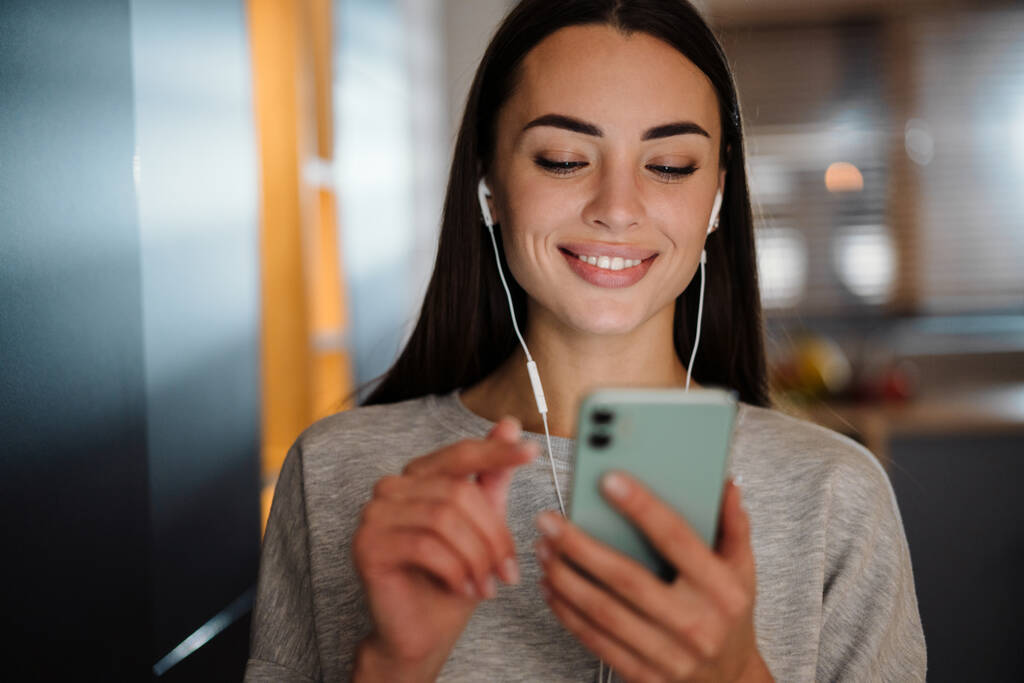 Улыбающаяся молодая женщина слушает музыку с наушниками и мобильным телефоном дома - Фото, изображение