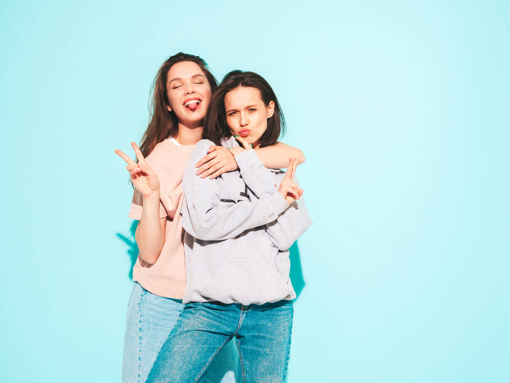 Deux jeunes belles femmes hipster souriantes en t-shirt blanc tendance d'été et des vêtements en jean. Femmes exy insouciantes posant près du mur bleu clair en studio.Modèles positifs - Photo, image