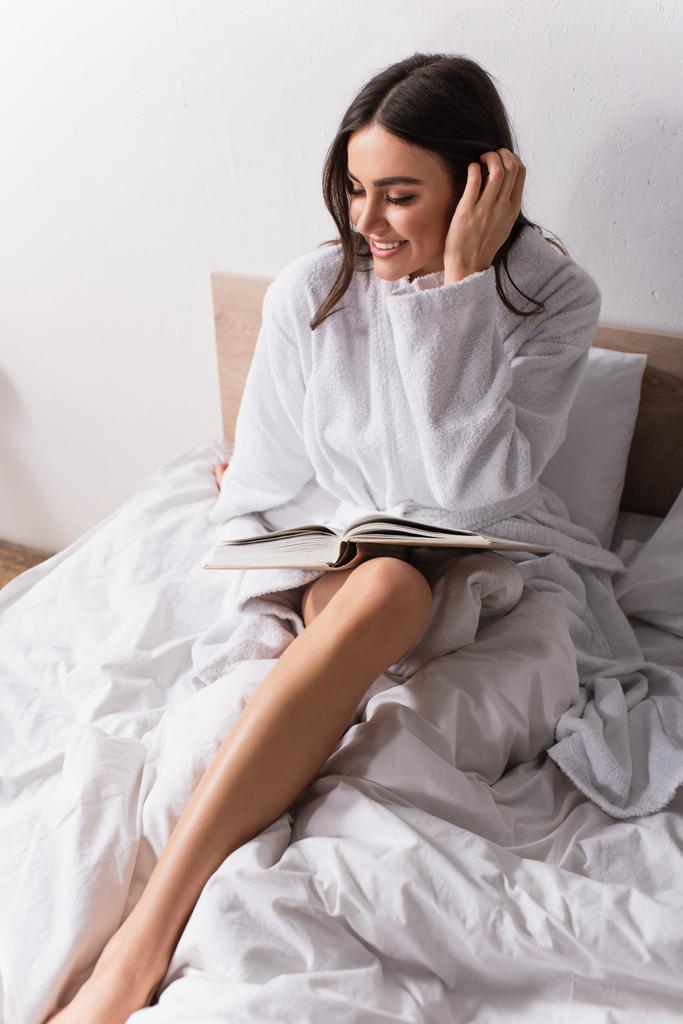 χαμογελαστή γυναίκα προσαρμόζει τα μαλλιά, ενώ διαβάζοντας μυθιστόρημα στην κρεβατοκάμαρα  - Φωτογραφία, εικόνα