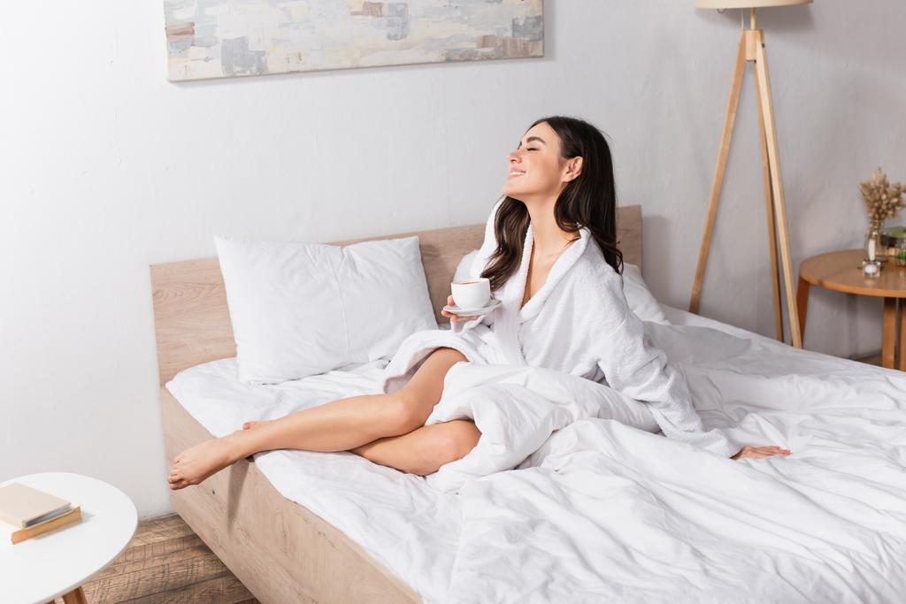 boldog fiatal nő kezében csésze kávét és csészealj a hálószobában  - Fotó, kép