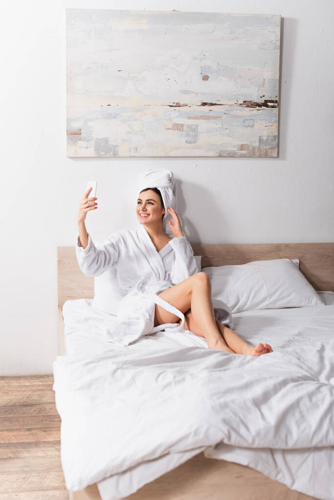 счастливая женщина в полотенце на голове и халате делает селфи в спальне  - Фото, изображение