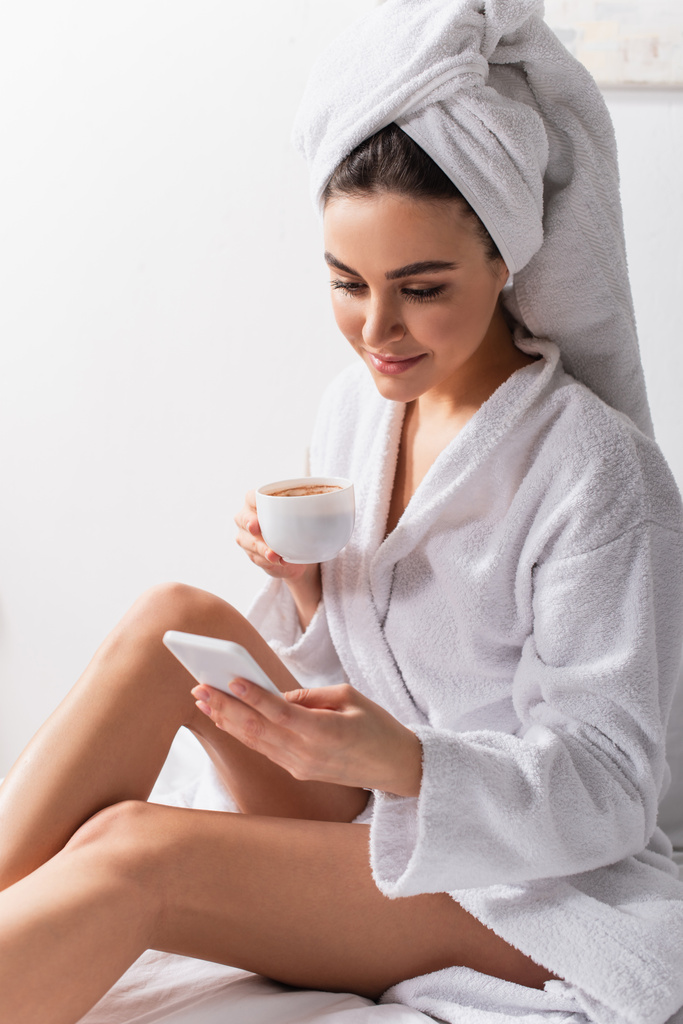 donna felice in asciugamano sulla testa e accappatoio utilizzando smartphone e tenendo in mano una tazza di caffè in camera da letto  - Foto, immagini