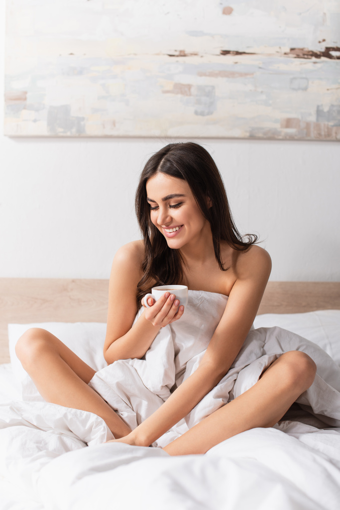 young brunette woman smiling while holding cup of coffee in bed - Photo, Image