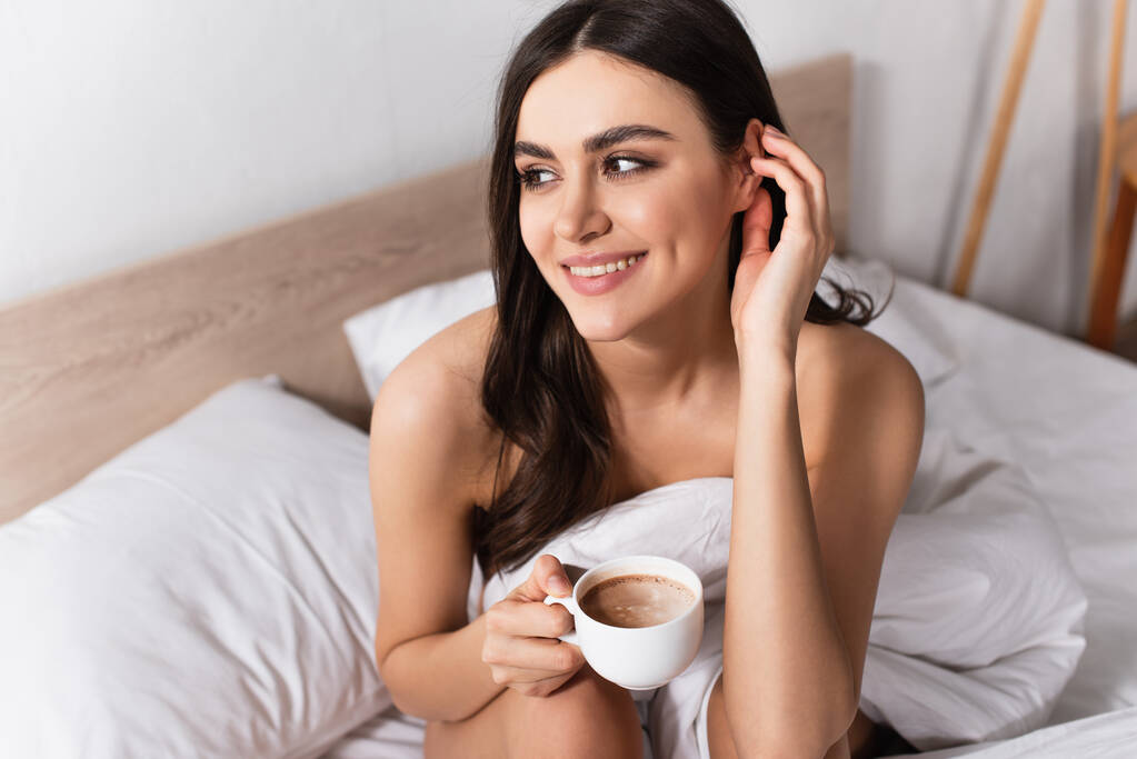 brünette Frau lächelt, während sie ihre Haare verstellt und eine Tasse Kaffee im Bett hält - Foto, Bild