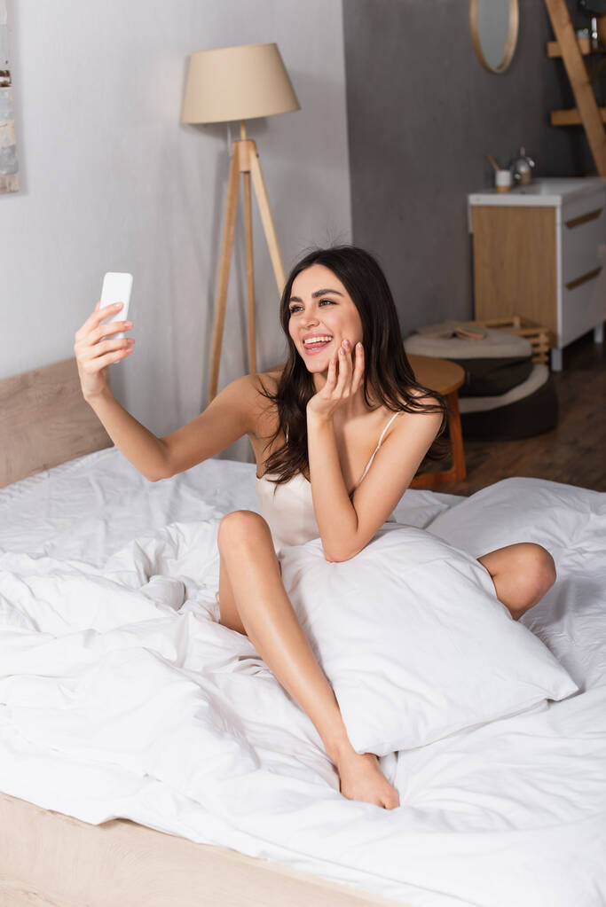 alegre joven mujer sobresaliendo lengua y tomando selfie mientras está sentado en la cama - Foto, Imagen