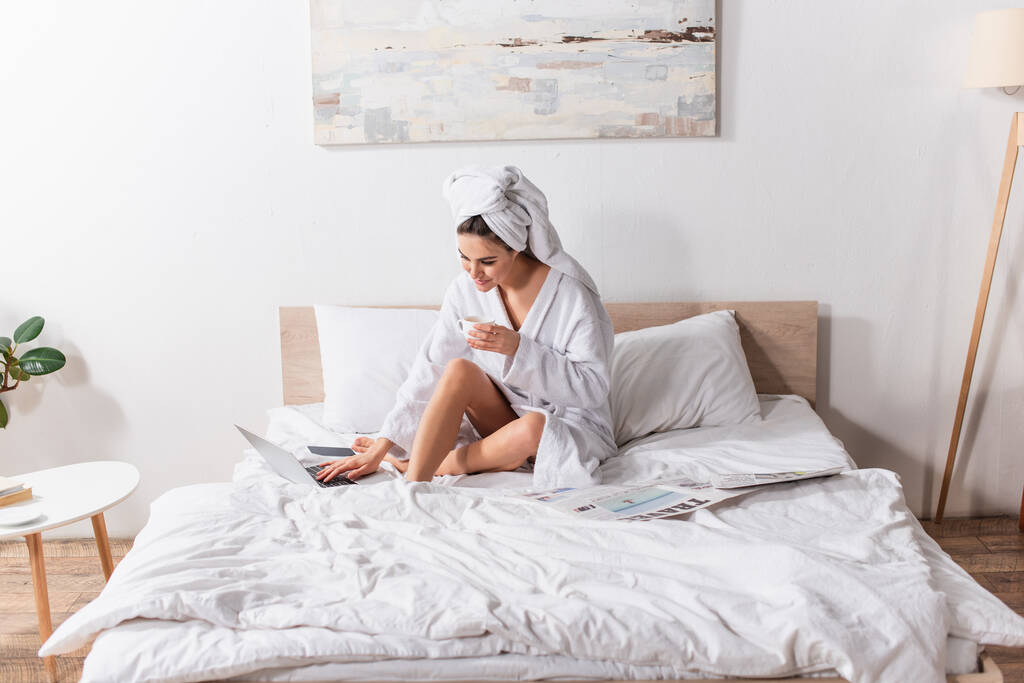 χαρούμενη γυναίκα με μπουρνούζι και πετσέτα στο κεφάλι κρατώντας φλιτζάνι καφέ και χρησιμοποιώντας φορητό υπολογιστή κοντά σε smartphone και εφημερίδα στο κρεβάτι  - Φωτογραφία, εικόνα
