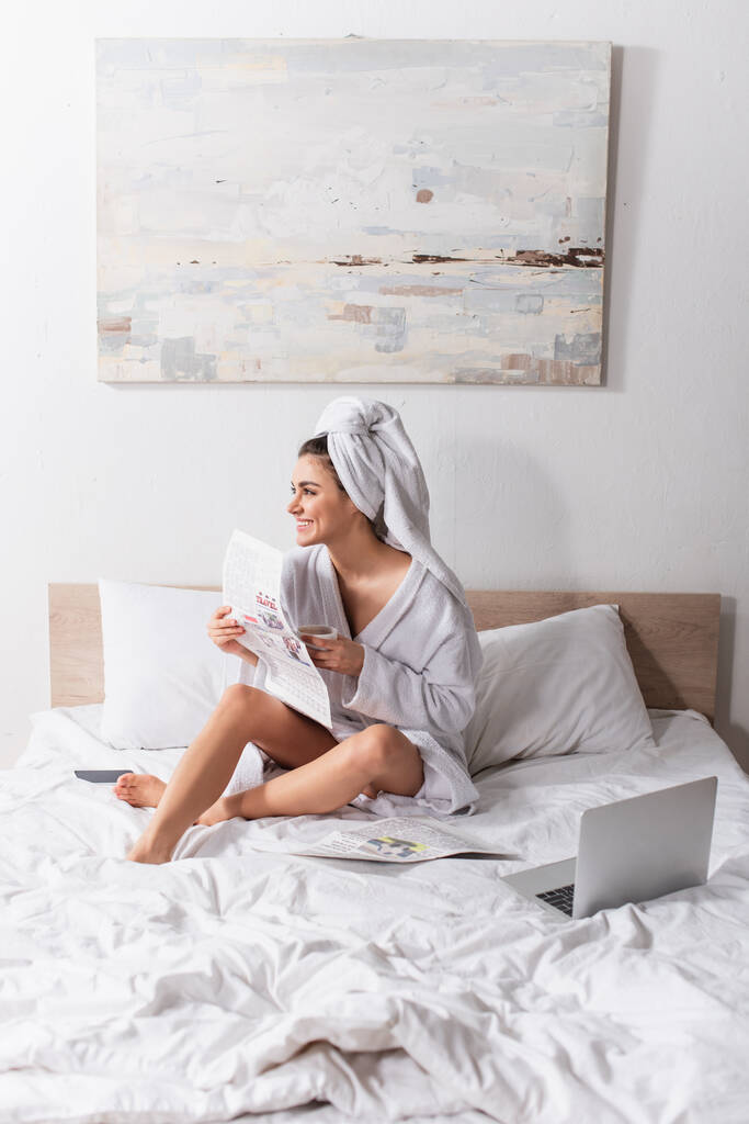 幸せな女性ですバスローブとタオル頭の上にコーヒーと新聞のカップを持っていますベッドの上のガジェットの近く  - 写真・画像