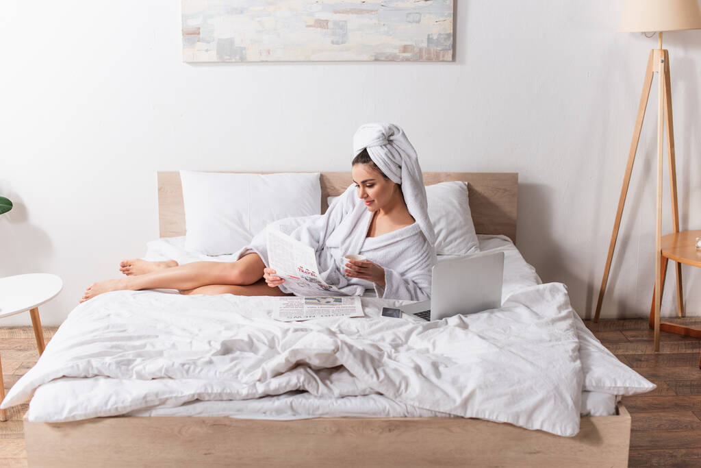 mujer joven en albornoz y toalla en la cabeza sosteniendo taza de café y periódico cerca de gadgets en la cama  - Foto, Imagen