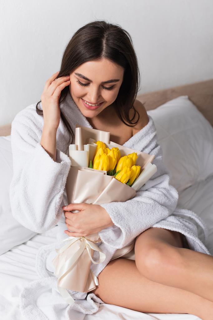 gelukkig vrouw in badjas houden boeket van gele tulpen in slaapkamer - Foto, afbeelding