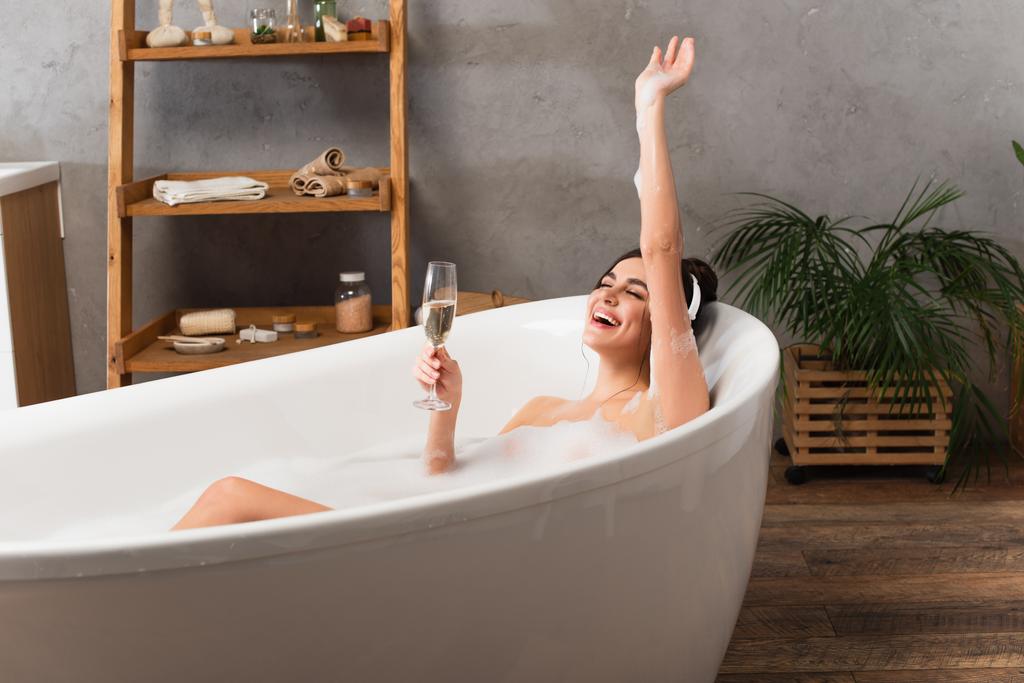 szczęśliwa kobieta w słuchawkach słuchając muzyki i trzymając kieliszek szampana podczas kąpieli  - Zdjęcie, obraz