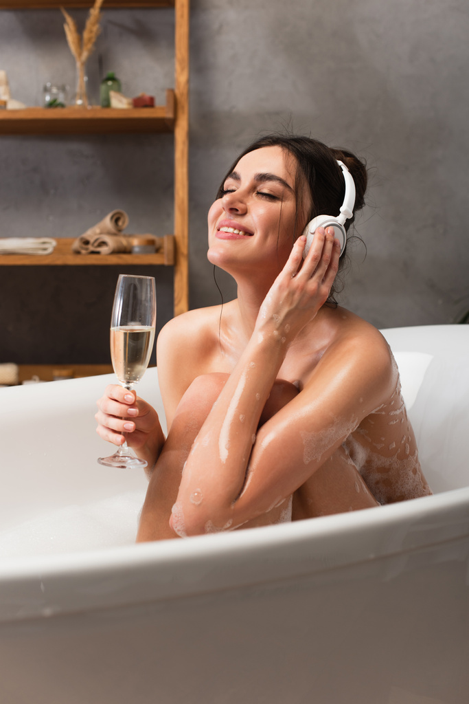 радостная женщина в наушниках слушает музыку и держит бокал шампанского во время принятия ванны  - Фото, изображение
