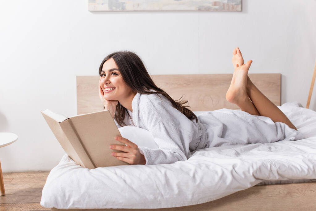 Χαρούμενη γυναίκα με μπουρνούζι ξαπλωμένη στο κρεβάτι και κρατώντας βιβλίο - Φωτογραφία, εικόνα
