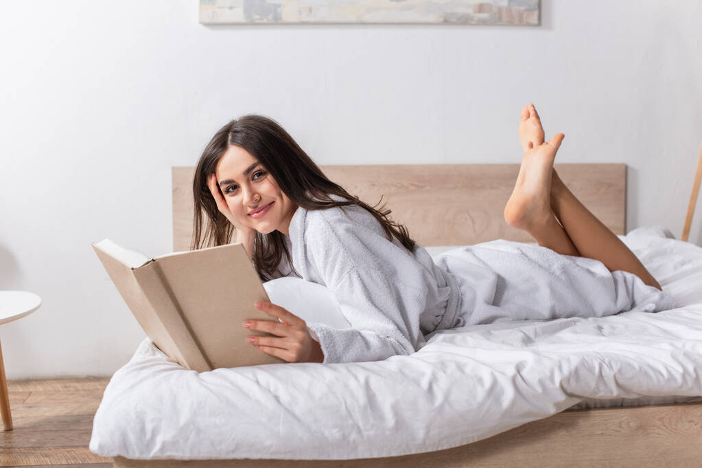 νεαρή χαρούμενη γυναίκα με μπουρνούζι ξαπλωμένη στο κρεβάτι και κρατώντας βιβλίο - Φωτογραφία, εικόνα