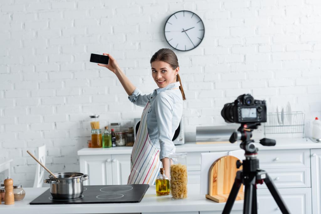 sonriente ama de casa tomando selfie cerca de la estufa y borrosa cámara digital en la cocina - Foto, imagen