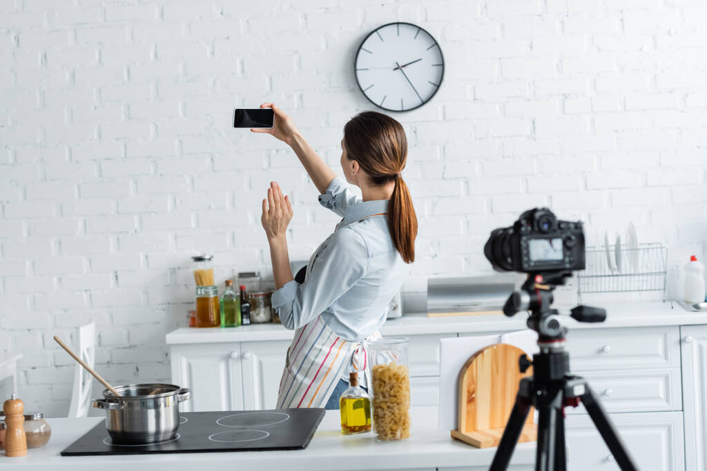 молодой кулинарный блоггер машет рукой возле смартфона с чистым экраном и размытой цифровой камерой на кухне - Фото, изображение