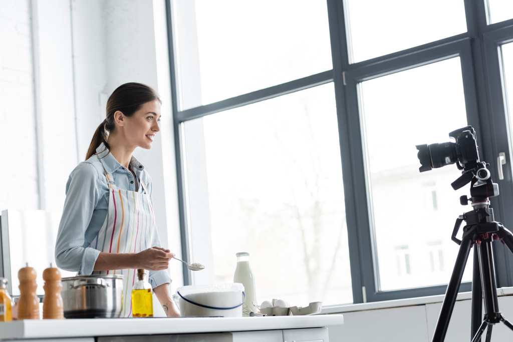 улыбающийся кулинарный блоггер держит ложку с мукой во время онлайн приготовления пищи перед цифровой камерой - Фото, изображение