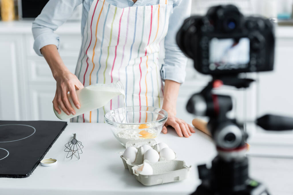 vue recadrée de la femme dans un tablier ajoutant du lait dans un bol avec de la farine et des œufs près d'un appareil photo numérique flou - Photo, image