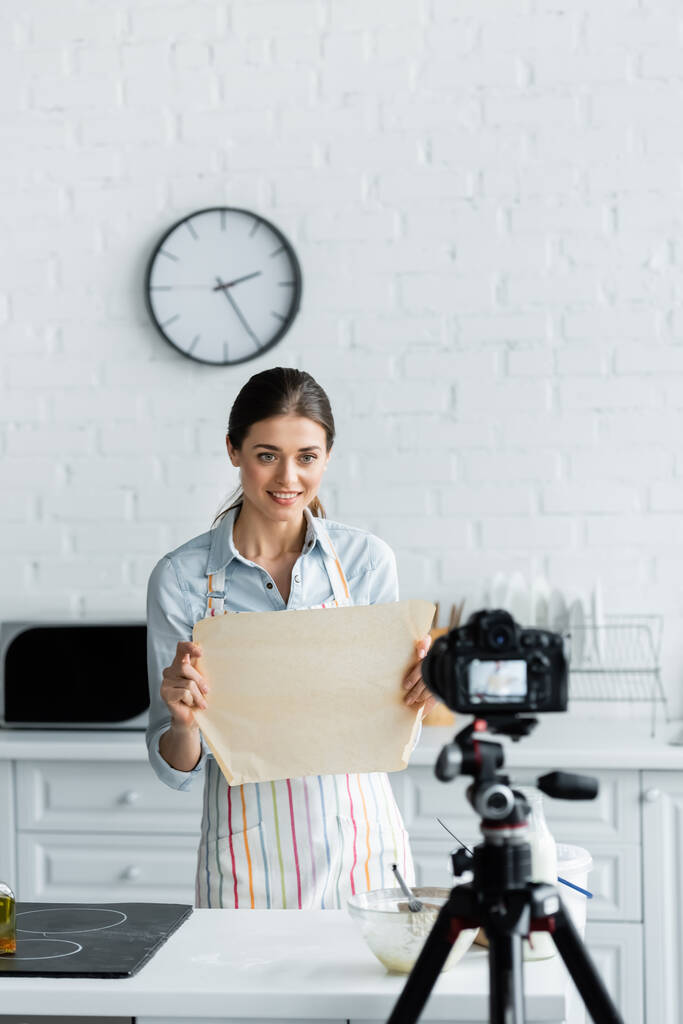 χαμογελαστός blogger μαγειρικής δείχνει περγαμηνή ψησίματος σε θολή ψηφιακή κάμερα στην κουζίνα - Φωτογραφία, εικόνα