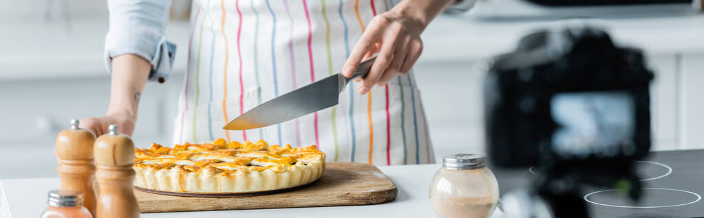 visão parcial do blogueiro culinário segurando faca perto de torta saborosa e câmera digital borrada, banner - Foto, Imagem