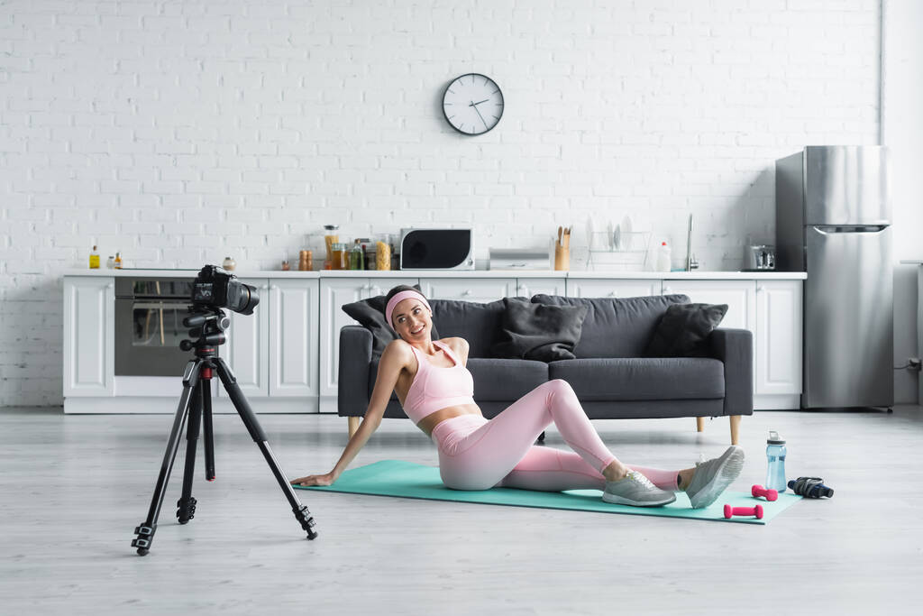 glimlachende vrouw in sportkleding trainen op fitness mat in de buurt van digitale camera - Foto, afbeelding