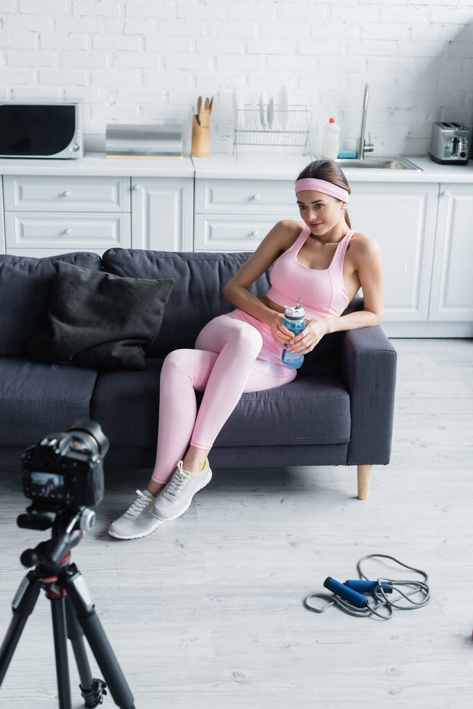 mulher com garrafa de esportes sentado no sofá perto da câmera digital no tripé - Foto, Imagem