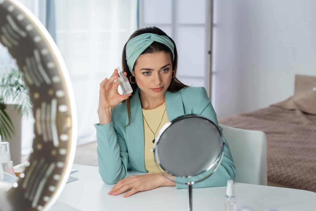 стильная женщина предлагает сыворотку для лица возле кольцевой лампы и зеркала - Фото, изображение
