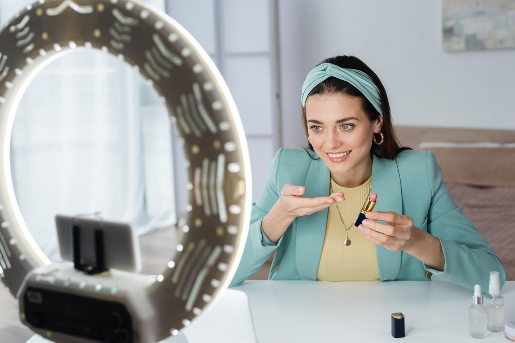 smiling woman pointing at lipstick near smartphone in holder with circle lamp - Photo, Image