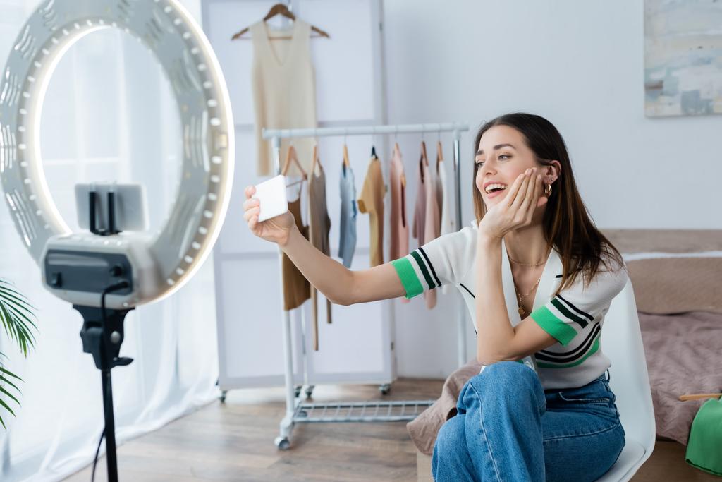 blogueur de mode heureux de prendre selfie près des cintres avec des vêtements et porte-téléphone avec lampe annulaire - Photo, image
